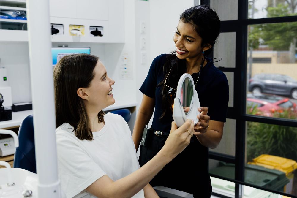 A woman smiles while holding a mirror, showcasing her new smile after a full mouth reconstruction at Northside Dental Solutions.